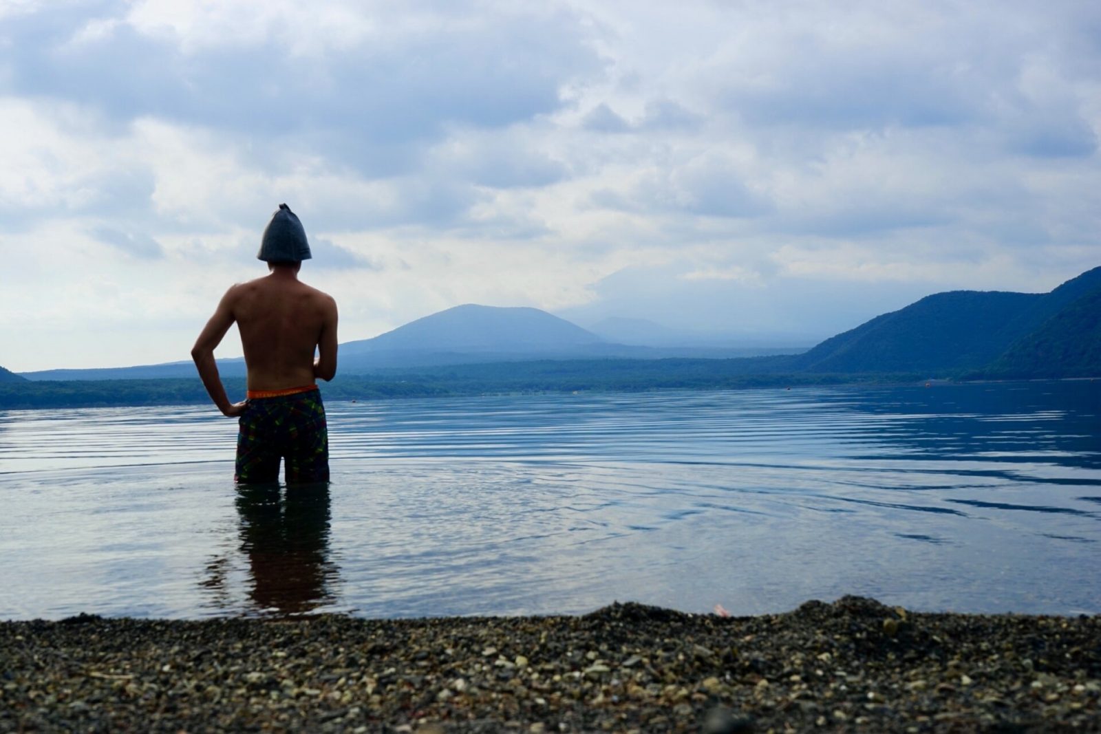 湖の中に立って遠方の山を眺める、サウナハットをかぶった男の人