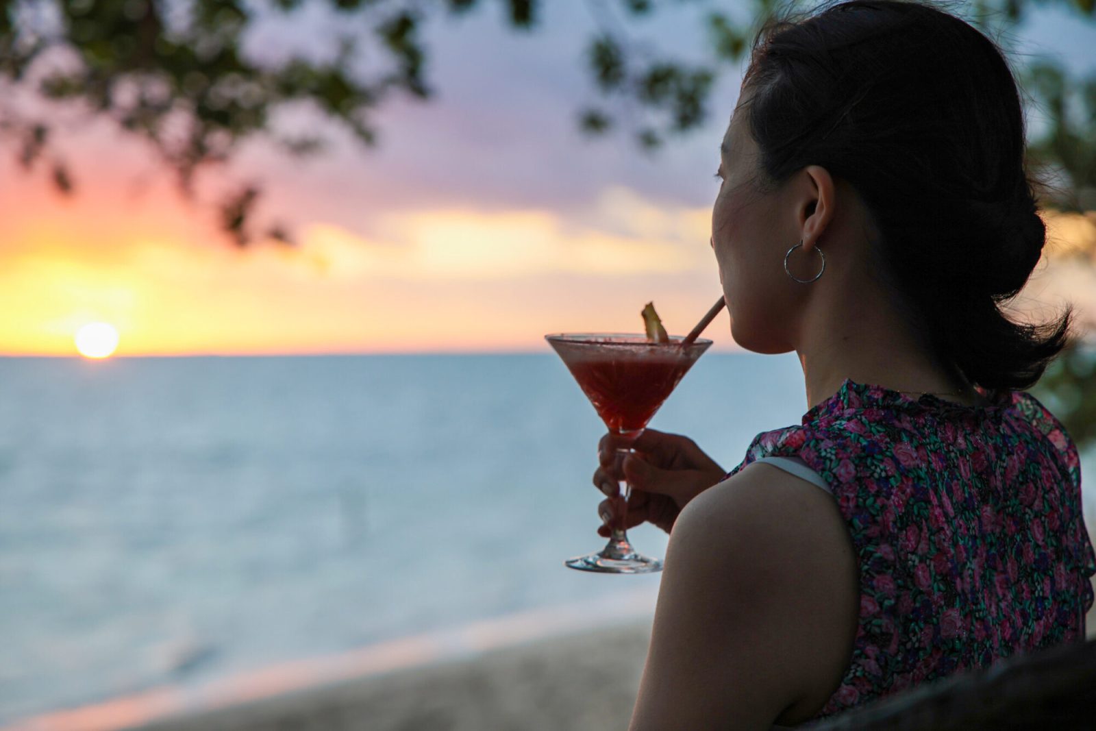 海に沈む夕日を見ている、片手でカクテルを持つ女性の姿