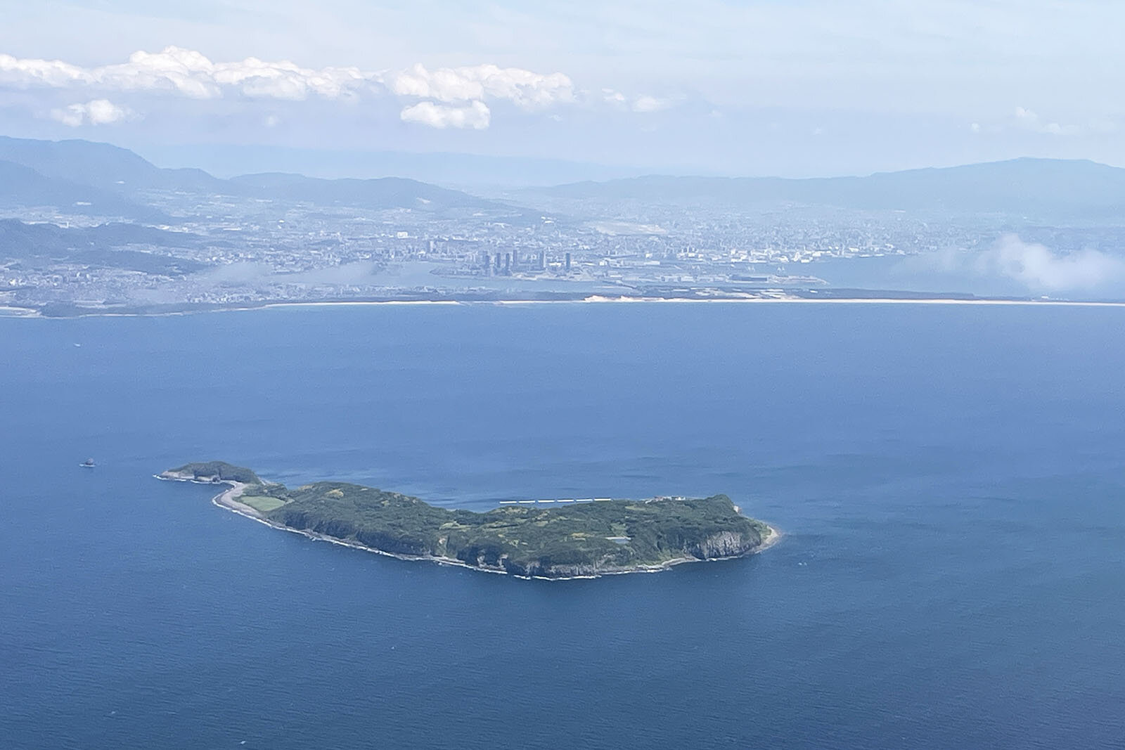 空撮で見下ろす、青く澄んだ海にぽつりと浮かぶ島の全貌と向こうに写っている山々が連なっている景色