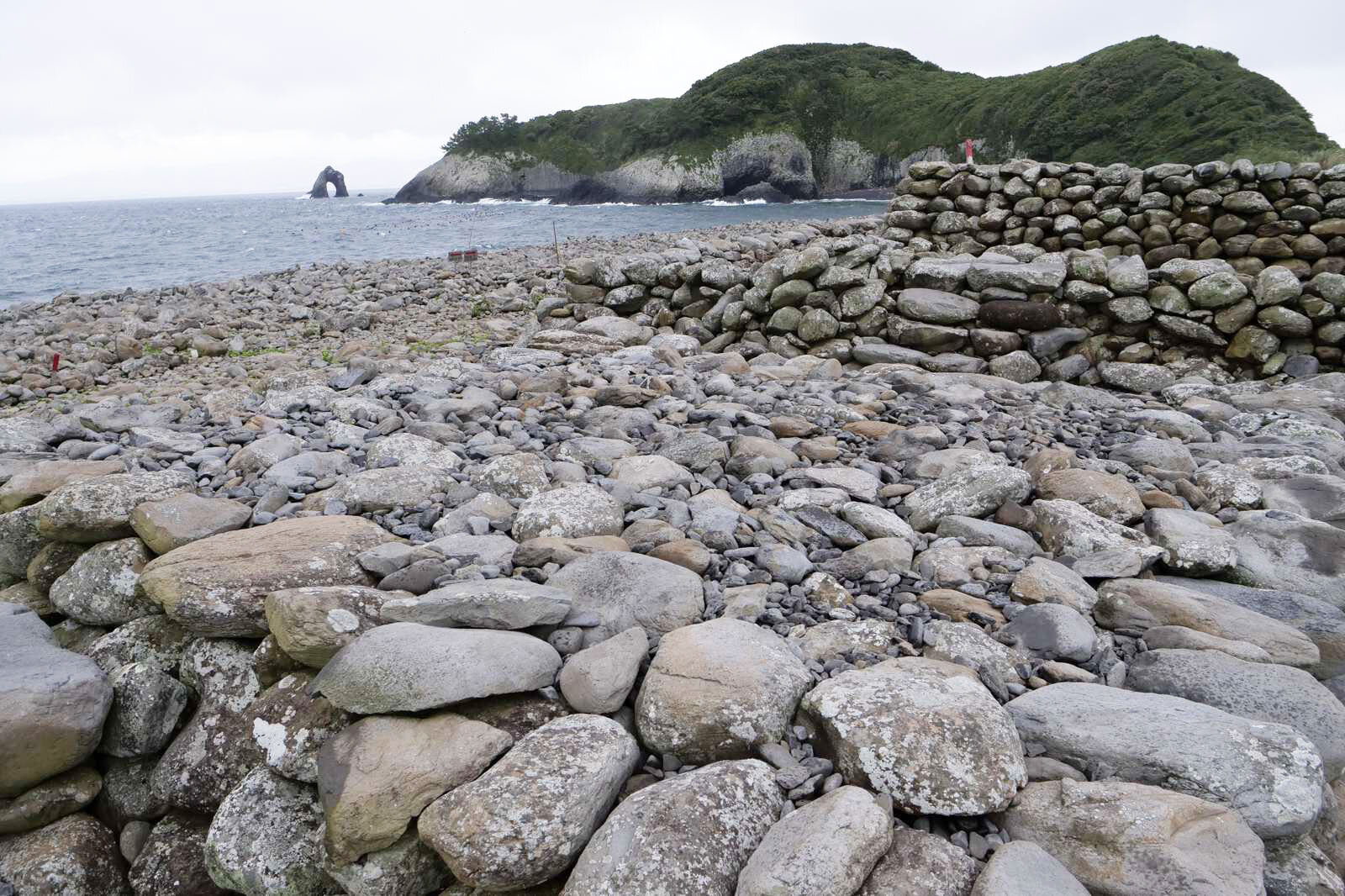 海辺に大きな石塚が広がっている。向こうには緑に覆われた岩壁と、海に立つ岩が見える
