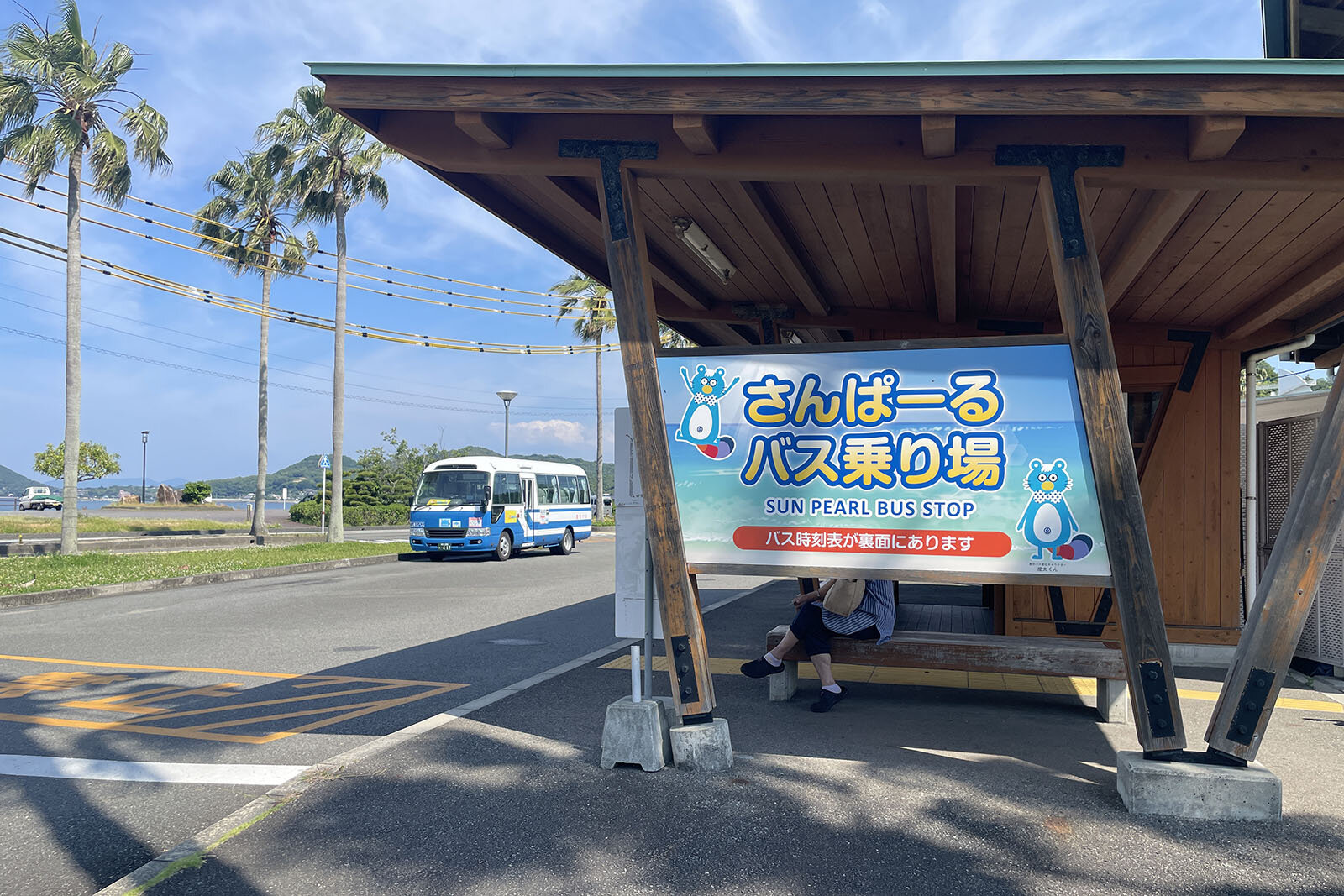 木で建てられた「さんぽーるバス乗り場」の看板の裏側に、人がひとり静かに座ってバスを待っている。斜め向かいにはバス一台が停まっている