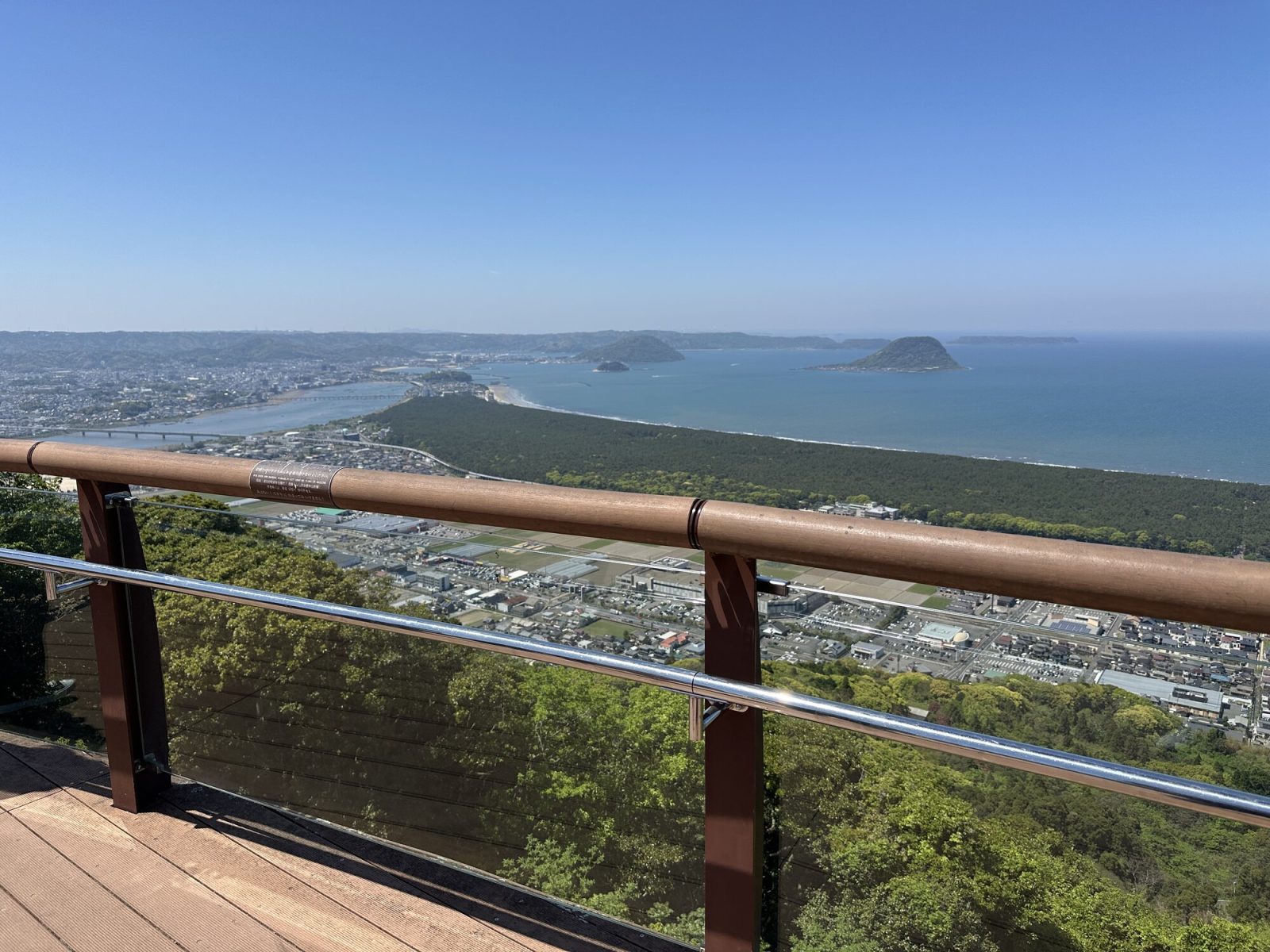 高い展望台から見た景色。海と緑の森、町並みが広がり、遠くに小さな島々が見える美しい風景。空は青く、よく晴れています